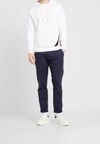 Witte hoodie met trekkoorden en zijaccenten in marineblauw en rood, gecombineerd met marineblauwe broek en witte sneakers met marineblauwe details.