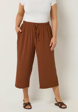 Frau trägt lockere braune, gekürzte Hose mit Kordelzugbund, weißes kurzärmeliges Oberteil und braune Sandalen, steht vor einem einfarbigen Hintergrund.