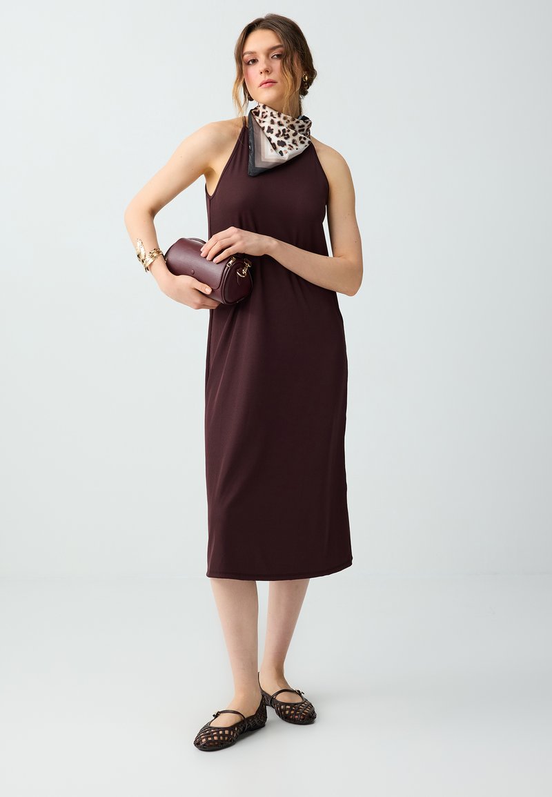Femme portant une robe midi sans manches violet foncé, foulard léopard au cou, tenant un petit sac à main bordeaux et portant des ballerines tissées noires.