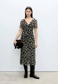 Schwarzes Blumenkleid mit kurzen, transparenten Ärmeln, V-Ausschnitt und fließender Silhouette. Kombiniert mit knielangen schwarzen Stiefeln und einer braunen Tasche.