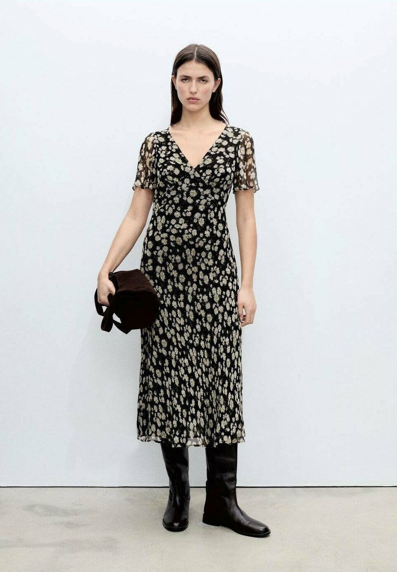 Schwarzes Blumenkleid mit kurzen, transparenten Ärmeln, V-Ausschnitt und fließender Silhouette. Kombiniert mit knielangen schwarzen Stiefeln und einer braunen Tasche.