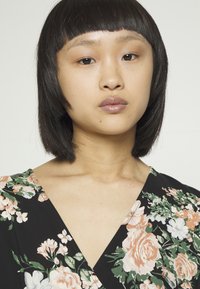 Tecido preto com padrão floral, apresentando flores em tons de rosa, branco e verde. Design em V. Textura suave. Modelo com cabelo preto liso em corte bob.