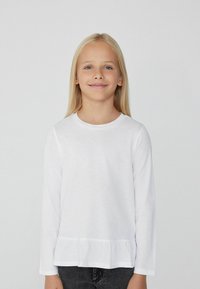 Biała koszula z długim rękawem, okrągłym dekoltem i falbanii na dole, wykonana z miękkiego materiału. Koszula ma luźny krój.