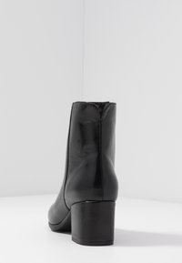 Botín de cuero negro hasta el tobillo con punta redonda y un tacón de bloque de 4 cm. Textura suave y detalle de costura mínima. Cremallera lateral no visible.