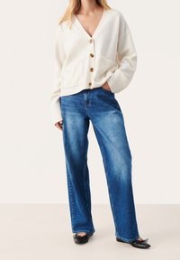 Witte gebreide cardigan met knoopsluiting en twee voorzakken, gecombineerd met blauwe denim jeans met wijde pijpen. Platte zwarte schoenen maken de outfit compleet.
