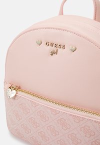 Sac à dos en simili cuir rose avec un dessus arrondi, doté de quincaillerie dorée, d'un logo et d'accents en forme de cœur, ainsi qu'une section inférieure à motif. Détails de zip dorés.