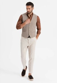 Mann in beigem Weste und Hose, braunem Hemd mit hochgekrempelten Ärmeln und dunkelbraunen Slippers, der mit einer Hand in der Tasche geht.