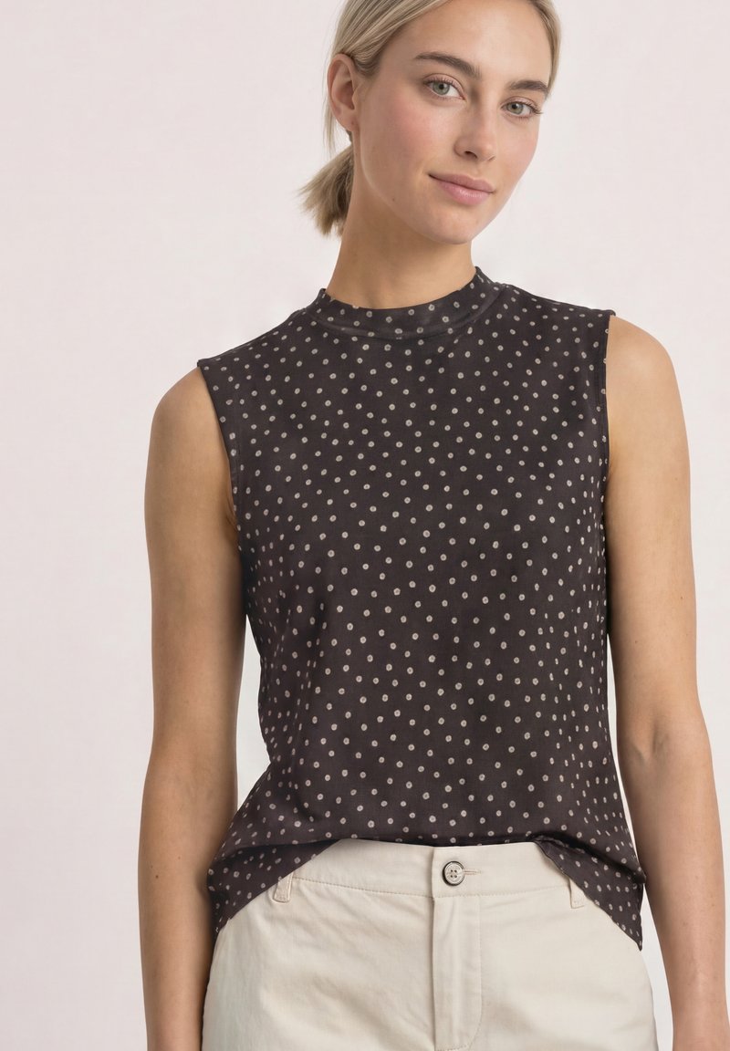 Femme aux cheveux blonds attachés en queue de cheval basse, portant un haut noir sans manches à pois blancs et un pantalon beige clair sur un fond uni.