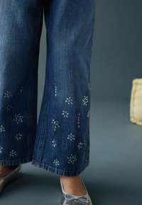Blugi din denim cu croială largă, de culoare albastru închis, cu broderie florală argintie lângă tiv. Asortați cu balerini argintii strălucitori.