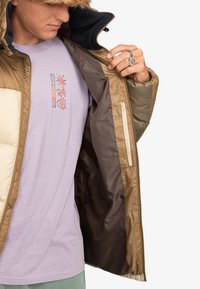 Jongere in een bruine pufferjack, die het openhoudt om een lila t-shirt met multicolore abstracte ontwerpen en tekst te onthullen.