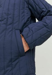 Marinblått quiltat jacka med ståkrage, sidofickor och en mjuk textur. Synlig sömnad löper vertikalt längs tyget.