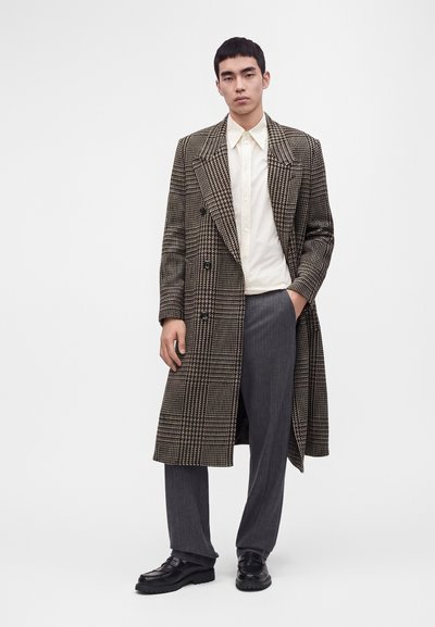 Homme debout portant un long manteau à carreaux, une chemise crème boutonnée, un pantalon gris et des chaussures noires, une main dans la poche, sur un fond uni.