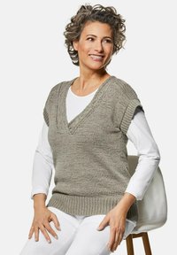 Lichtgrijs gebreid vest met een V-hals, korte opgerolde mouwen en een geribde zoom, gedragen over een wit shirt met lange mouwen. Glad textuur.