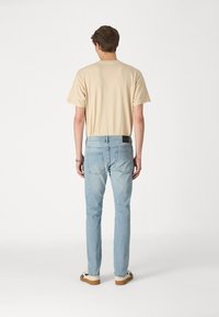 Beige t-shirt with a relaxed fit, paired with light blue slim-fit jeans. Visible stitching and back pockets detail. Brown-soled shoes.