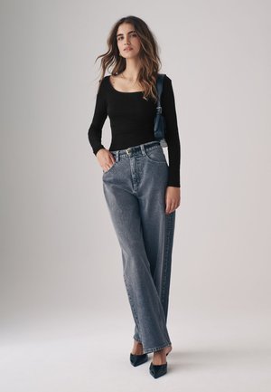 Vrouw staat en draagt een zwarte top met lange mouwen, hoge wijde jeans, zwarte puntige hakken en een donkerblauwe schoudertas op een egale achtergrond.