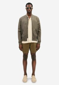 Olive green bomber jacket over a cream short-sleeve shirt, paired with brown shorts and beige slip-on shoes. Accessories include sunglasses.