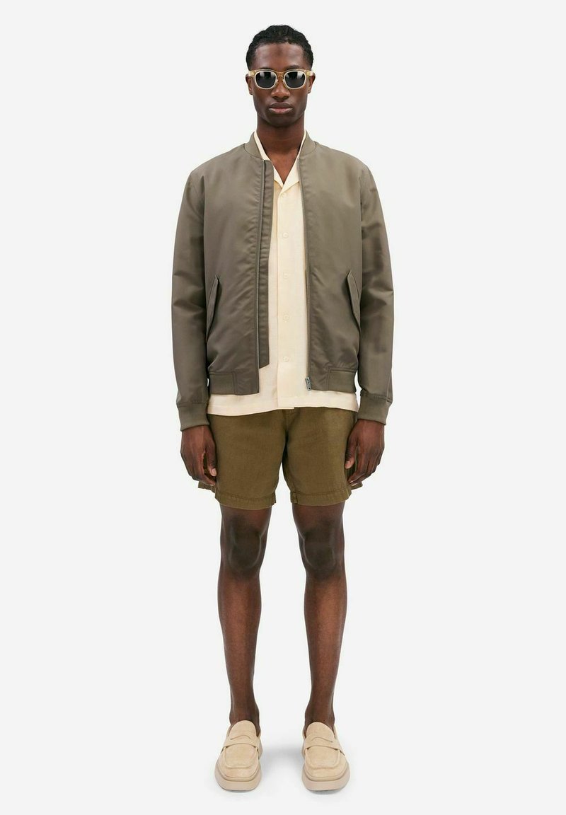 Olive green bomber jacket over a cream short-sleeve shirt, paired with brown shorts and beige slip-on shoes. Accessories include sunglasses.
