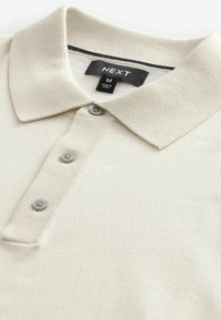 Polo de color beige claro hecho de tela texturizada, con cuello tipo polo, tres botones y una etiqueta de marca en el interior.