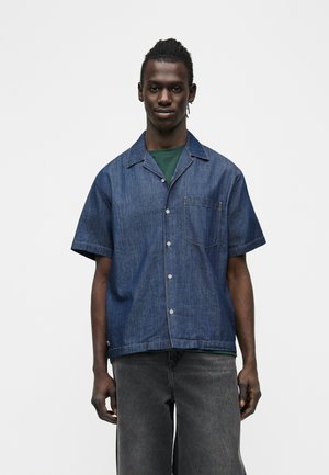 Giovane con capelli intrecciati che indossa una camicia di jeans a maniche corte sopra una T-shirt verde e jeans neri sbiaditi davanti a uno sfondo semplice.