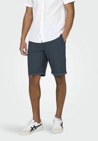 Shorts pour homme bleu foncé avec une surface texturée, ourlets roulés, poches latérales, associés à une chemise blanche à boutons et des baskets blanches.