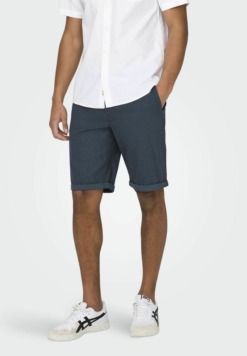 Shorts pour homme bleu foncé avec une surface texturée, ourlets roulés, poches latérales, associés à une chemise blanche à boutons et des baskets blanches.