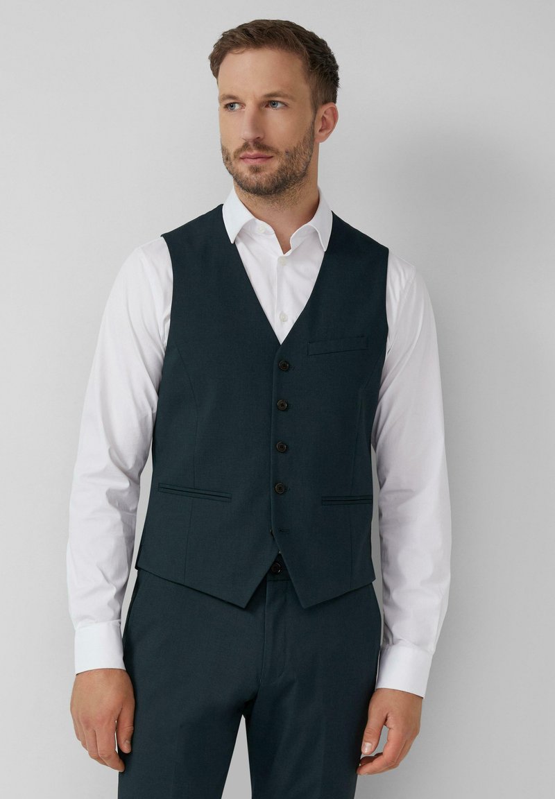 Man in een donkerblauw knoopvest over een wit dress shirt, met bijpassende donkerblauwe broek, staande tegen een effen lichte achtergrond.
