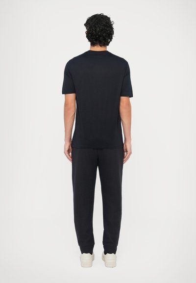 Čierne bavlnené tričko s krátkymi rukávmi, v kombinácii s čiernymi textúrovanými teplákmi a bielou obuvou, fotografované zozadu.