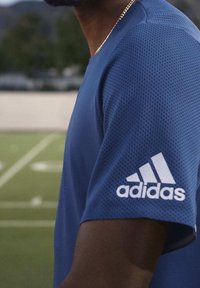 Modrá sportovní košile z texturované, prodyšné látky. Na levém rukávu se nachází bílé logo Adidas. Standardní střih, krátké rukávy.