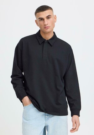 Junger Mann mit kurzen Haaren, der ein schwarzes Langarm-Poloshirt und hellblaue Jeans trägt, steht mit einer Hand in der Tasche.