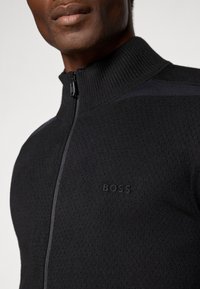 Sweater preto com fecho de correr e gola alta, apresentando um padrão texturizado e uma marca discreta ao centro. Feito de um tecido de malha.