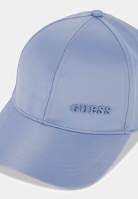Gorra de béisbol azul clara con visera curva y logo en relieve "GUESS" en el panel frontal, mostrada sobre un fondo blanco.