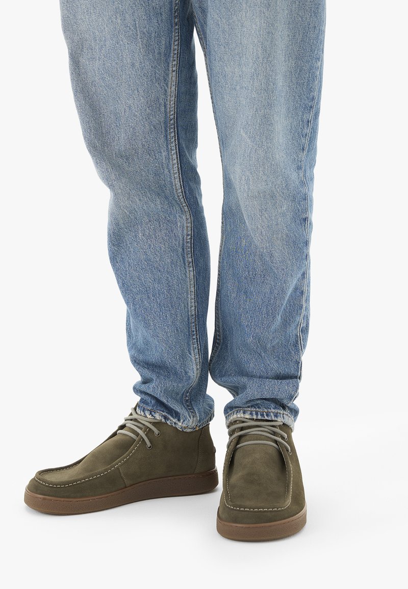 Personne portant un jean bleu clair et des chaussures en daim vert olive à lacets, debout sur une surface blanche.