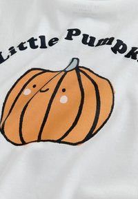 Cămașă albă din bumbac cu un grafic de dovleac portocalie, cu contururi negre, detalii faciale zâmbitoare și textul "Little Pumpkin."