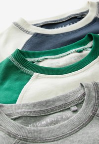 Trois t-shirts à col rond pliés en gris, vert et blanc, et bleu marine et blanc, avec des coutures visibles et des étiquettes de vêtements de nuit.