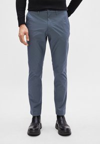 Schlanke, mittelhohe Hose aus hellblauem Stoff mit glatter Textur, mit Reißverschluss, vorderen Taschen und getragen mit schwarzen Knöchelstiefeln.