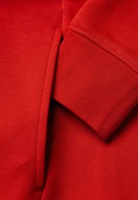 Detailní záběr na červenou mikinu s kapucí, konkrétně na kapsu a rukáv, který ukazuje podrobné švy a pletenou texturu.
