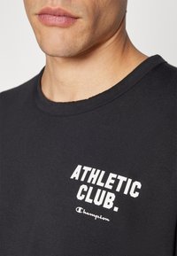 T-shirt en coton noir avec un col rond et un texte blanc imprimé "ATHLETIC CLUB." avec le logo "Champion" en dessous. Texture lisse, design décontracté.