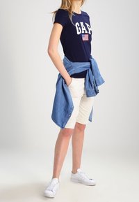 Námorní t-shirt s logem "GAP" a vlajkou, doplněný béžovými šortkami a lehkou džínsovou košilí uvázanou v pase. Bílé tenisky dokončují vzhled.