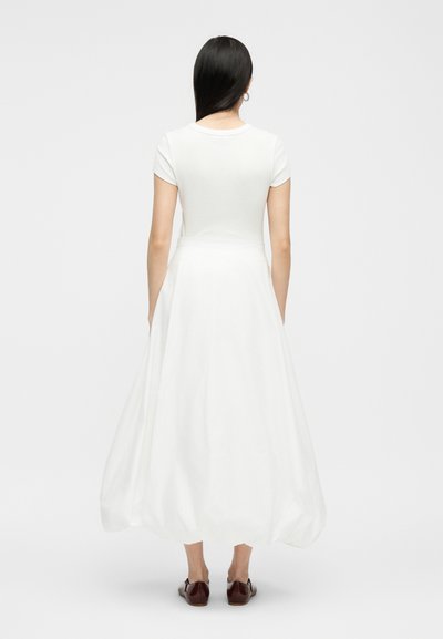 Γυναίκα με μακριά μαύρα μαλλιά, φορώντας ένα λευκό μίντι φόρεμα με κοντά μανίκια και καφέ επίπεδα παπούτσια, στέκεται γυρισμένη με την πλάτη σε απλό φόντο.