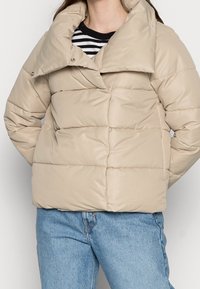 Beżowa kurtka puchowa z wysokim kołnierzem i zapięciem na zatrzaski, o pikowanym wzorze. Stylizowana z paskowaną koszulą pod spodem i niebieskimi dżinsami.