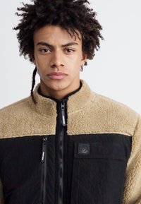 Fleecejacke mit einem oberen Teil aus braunem Sherpa und unteren Abschnitten aus schwarzem Stoff. Verfügt über Frontreißverschlüsse und ein Logo-Patch auf der Brust.