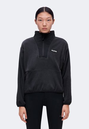 Person iført en sort Columbia fleece-pullover med høj krave og lynlås foran, stående mod en ensfarvet lys baggrund.