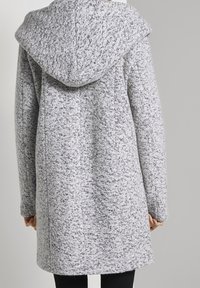 Šedý strukturovaný kabát s velkou kapucí, který má skvrnitý vzor, rovný střih a dlouhé rukávy. Materiál vypadá jemně a teple.
