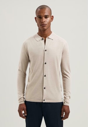 Beige blouse met lange mouwen en een knoopsluiting, zachte textuur en een kraag, gecombineerd met marineblauwe onderkleding. Heeft vijf zwarte knopen.