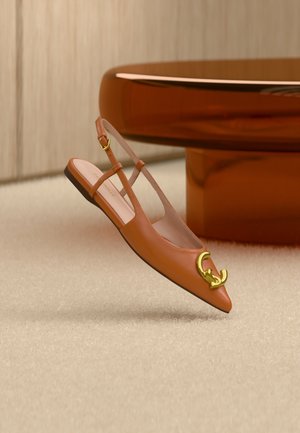 Bruine leren slingback platte schoen met puntige neus en gouden verstrengeld C-logo op een beige tapijt nabij een bruine tafel.