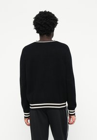Sort strikket sweater med rund hals, der har hvide striber ved kanten og manchetterne. Designet er løstsiddende med lange ærmer.