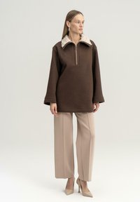 Pullover in pile marrone con collo zip, colletto testurizzato crema, maniche ampie, abbinato a pantaloni ampi beige e tacchi beige.