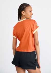 T-shirt en coton orange à manches courtes avec des accents crème, design court, et détail de logo sur la manche, associé à un short noir.