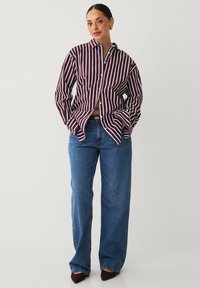 Striped cotton shirt in burgundy and white, buttoned up, paired with high-waisted blue denim jeans. Model stands with hands in pockets.
