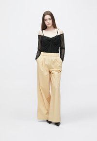 Svart långärmad mesh-topp med vriden detalj framtill, kombinerad med högmidjade, vida beige byxor. Modellen står med händerna i fickorna.